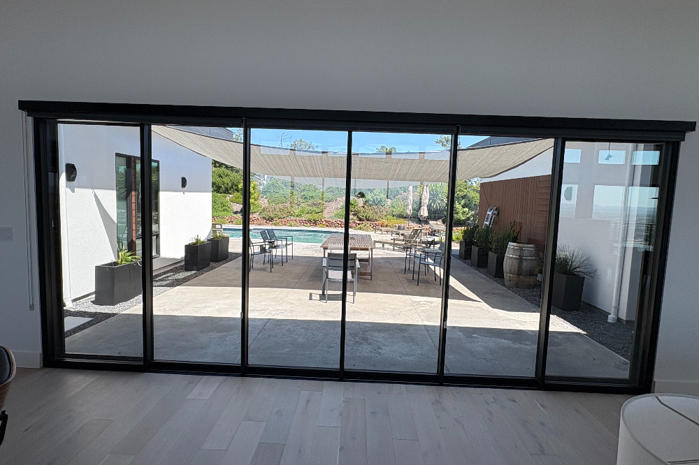 Sliding glass doors open to a sunlit outdoor patio with a pool, shade sails, and dining tables.