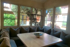 IMG_4063 - Transparent Window Washing Sunlit corner dining area with a light wood table, gray cushioned U-shaped seating, and a woven circular pendant light overhead.