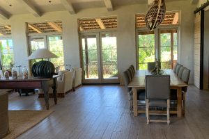IMG_4066 - Transparent Window Washing Open-plan dining room with a long wooden table and gray upholstered chairs, large glass doors to a green garden, and natural light flooding the space under exposed beams.