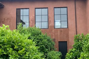 IMG_5275 - Transparent Window Washing Brown stucco building with three tall square windows and a dark door, partially obscured by dense green shrubs.