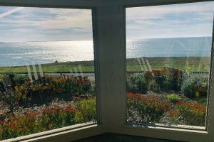 Cozy bay window view of a sunlit ocean and colorful flower garden outside the window.