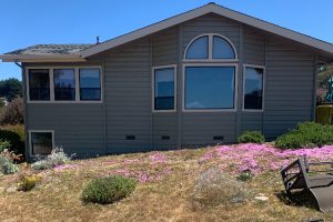 Gray suburban house with a large central arched window, surrounded by pink flowering ground cover in the yard.