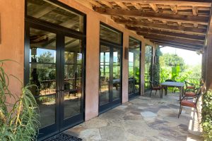 IMG_7066 - Transparent Window Washing Outdoor patio with glass doors along a stone-tiled floor under a wooden beam ceiling, with a table and chairs and surrounding greenery.