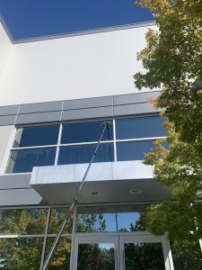 IMG_7354 - Transparent Window Washing Modern white building exterior with large reflective glass windows, a window-cleaning pole reaching the panes, blue sky above, and trees nearby.