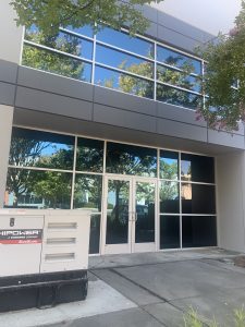 IMG_7419 - Transparent Window Washing Office building exterior with large glass windows reflecting trees and blue sky; foreground shows a gray utility box labeled 'HIPOWER' (generator).