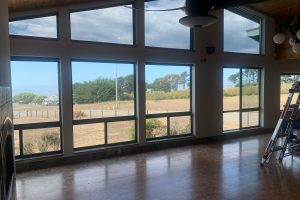 Interior room with tall windows looking out to a dry field and trees; a ladder leans against the right wall.