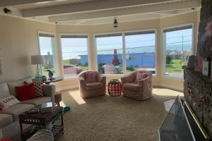 Bright living room with bay windows overlooking the ocean; two pink armchairs flank a red lattice side table beside a beige sofa with red cushions in front of a stone fireplace.