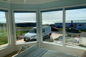 IMG_8070 - Transparent Window Washing Bright bedroom with blue bedding and a bay window view of a coastal street, a white van parked outside and the ocean visible beyond.