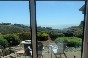 IMG_8165 - Transparent Window Washing Glass doors frame a sunny coastal garden with patio chairs and a circular fire pit, overlooking the ocean.