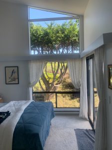 IMG_8169 - Transparent Window Washing Bright bedroom with a blue quilted bedspread, white curtains, and a large window showing green trees outside.
