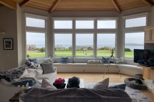 IMG_8675 - Transparent Window Washing Bright living room with a curved bay window overlooking the ocean; white sectional sofa, blue pillows, and a tripod camera set up near the window.