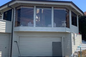 IMG_8797 - Transparent Window Washing Two-story house with a large bay window above a closed garage door, light gray siding and clear blue sky behind the property