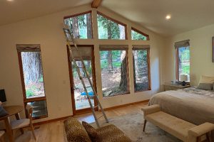 Bedroom with a ladder propped by a wall of tall, multi-pane windows overlooking a wooded yard; a bed with a bench sits to the right.