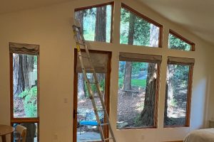 Ladder leaning inside a bright room with large wood-framed windows overlooking a wooded yard. 2) Interior renovation scene: tall windows and a metal ladder, with forest outside. 3) Partially finished living space with oversized windows and a propped interior ladder. 4) Sunlit room with triangular clerestory windows and a ladder, overlooking a patio and trees. 5) Open woodland view through multiple windows as a ladder rests inside a white-walled room.