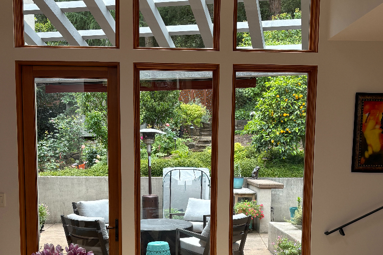 View through tall wooden-framed glass doors to a patio with seating and lush garden beyond.
