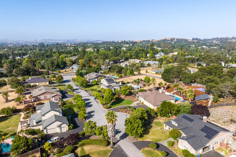 Aerial view of a sunlit suburban neighborhood with houses, green lawns, and tree-lined streets, seen from above.