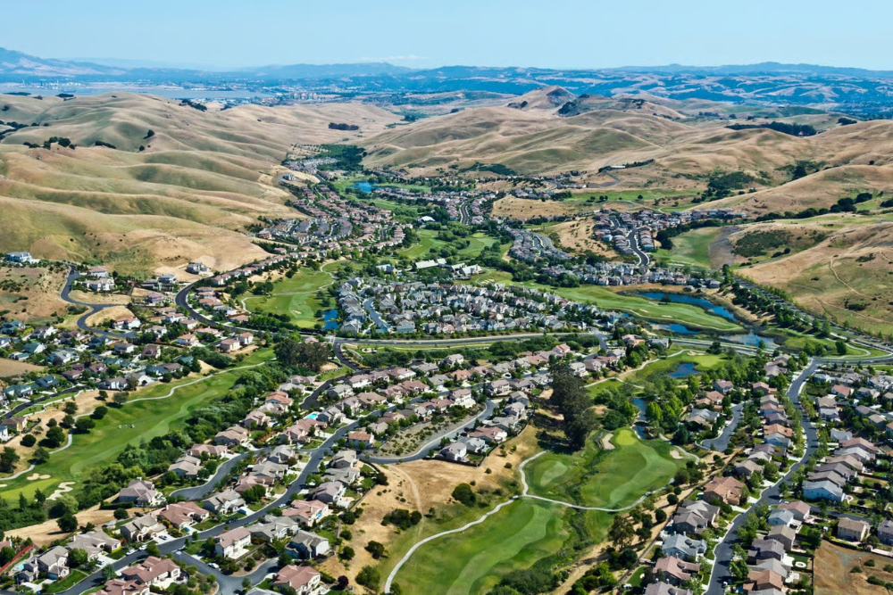 Aerial view of a suburban golf course community with winding roads, green fairways, and brown rolling hills in the distance.
