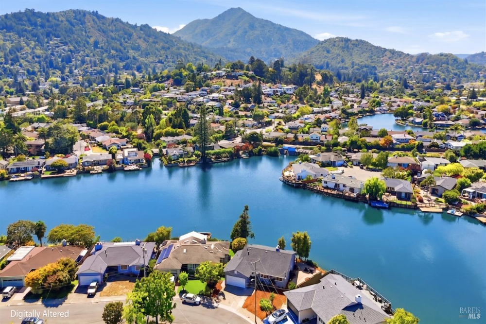 Aerial view of a suburban lake neighborhood with houses lining the water and green hills in the distance.