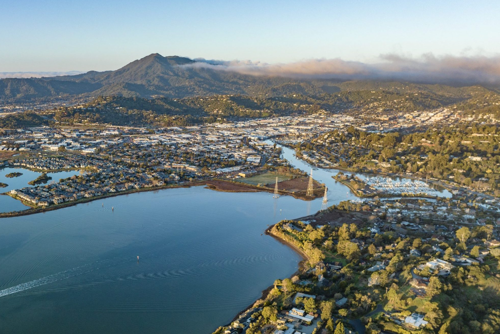 Aerial view of a lakeside town with a winding river, marina, and hills with a mountain backdrop under a clear sky.