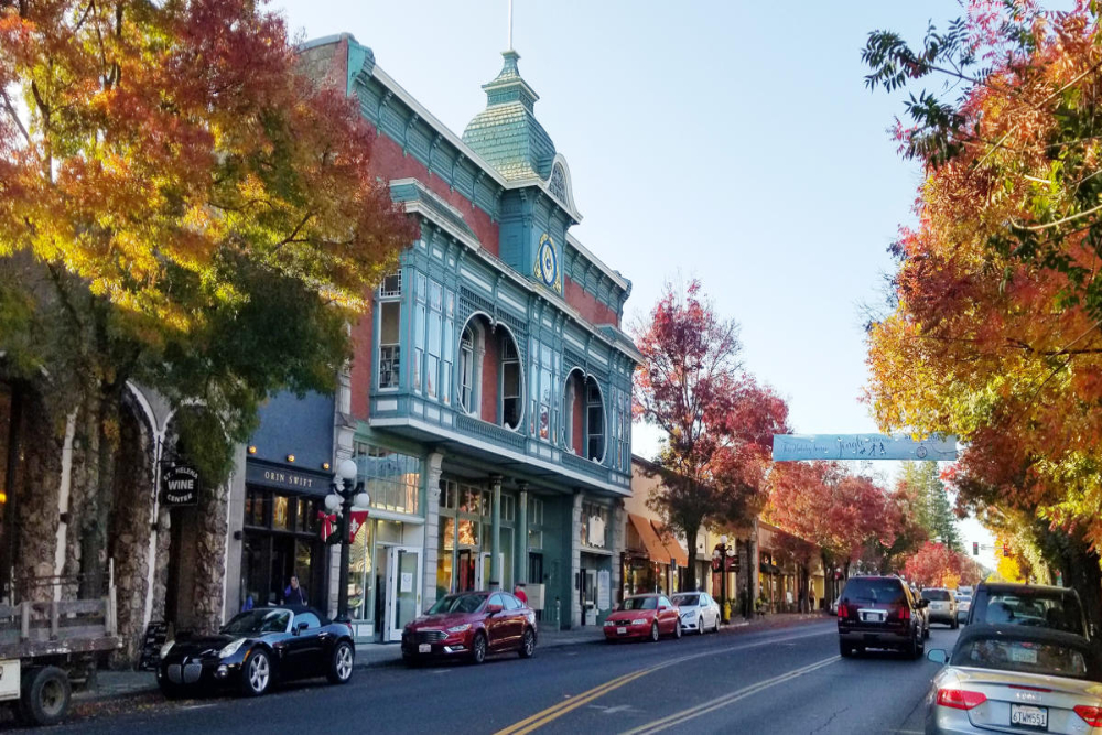 Autumn street scene with colorful trees lining a downtown block and a blue ornate two‑story storefront beside storefronts and parked cars.
